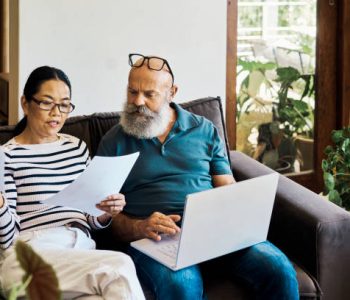 Senior interracial couple using a laptop and going retirement documents online on the couch at home. Mature husband and wife talking and reading paperwork while using a pc on the sofa in the lounge